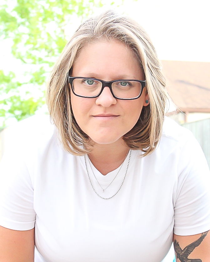 Photo of Ash sitting outside and looking at the camera. She has shoulder-length dirty blonde hair, glasses, a white t-shirt, and two necklaces.